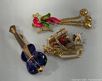 Photo showing all three brooches together: blue violin, gold/red ship, and colorful peacock
