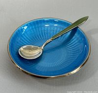 Small round sterling silver dish with vibrant blue guilloche enamel interior and silver rim, paired with a sterling silver spoon with green guilloche enamel handle placed inside the dish.