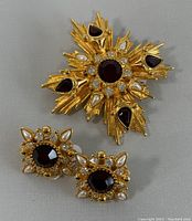 Lot showing the brooch and earrings on a gray background, top view showing Maltese cross design with red teardrop shaped and round rhinestones, faux pearls on gold tone.