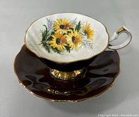 Angled view of the teacup showing the chocolate brown exterior and vibrant black-eyed Susan flower design on the interior with scalloped and gold-trimmed edges.