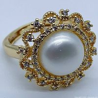Close-up angled view of ring showing white pearl center and surrounding clear stones on gold-plated decorative setting.