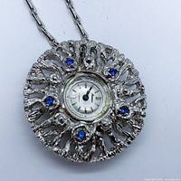 Close-up front view of the round textured pendant watch showing silver-tone case, blue stones, and watch face.