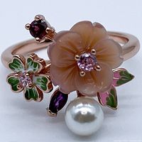 Front angled view showing carved peach flower with central pink gemstone, enamel leaves, rhodolite garnets, and pearl accent