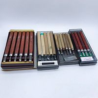 Four sets of wood carving tools displayed horizontally in trays. Handles are wooden in various colors, blades metal.