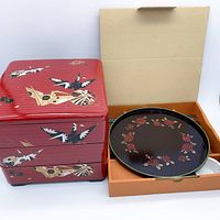 Full view of closed red three-tiered lacquer box decorated with origami cranes next to the boxed round sushi plate showing floral pattern.