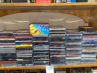 Five stacks of miscellaneous music CDs in jewel cases displayed on wooden shelves, showing spines with artist and album titles including The Beatles, Rod Stewart, U2, Bryan Adams, and others.