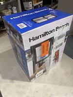 Box showing Hamilton Beach branding and image of the coffee maker with hand pressing a button, labels on box.