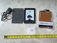 Photo of the Kindle Paperwhite with its grey folio cover opened, charging cord, and Incipio Antique Journal case in packaging, with measuring tape showing size.