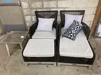 Photo of the full furniture set showing two chairs, two ottomans, cushions, and a wooden side table against a block wall