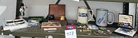 Full shelf view showing a collection of military memorabilia including mugs, boxes with cuff links and tie clips, badges, pins, and assorted items spread across a shelf.
