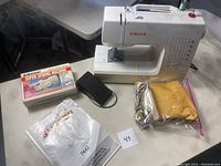 Singer 7442 sewing machine alongside foot pedal, instruction manual, cover, and an Emson handheld sewing machine in box on white table.