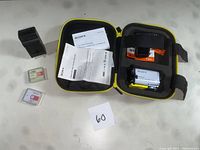 Full kit unboxed showing camera in waterproof housing, live-view remote, two spare batteries, wall charger, instruction manuals, in a black and yellow protective case.