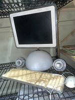 Front and angle view of 2002 Apple iMac all-in-one computer showing display, dome base, two clear dome speakers, keyboard and mouse on black shelving.