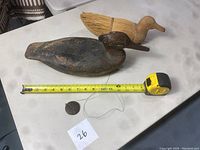 Two duck decoys with a tape measure for scale, showing that the wooden duck decoy is about 15 inches long and a straw duck decoy behind it.