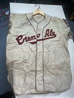 Full view of vintage cream baseball shirt with 'Cremo Ale' lettering and button-up front on hanger.