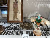 Photo showing framed Native American cross-stitched art, wooden Native American figure, two round engraved decorative copper plates, and several bolo ties on a shelf.