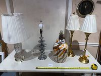 Five lamps displayed on a table including a large ceramic lamp with a plastic shade, two brass accent lamps with white pleated shades, a modern textured glass lamp, and a modern silver metal lamp base without shade.