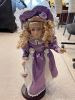 Porcelain doll standing on wooden base stand dressed in purple Victorian-style dress with lace and bonnet.