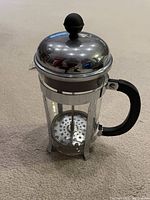 Photo showing French press coffee maker on carpeted floor, highlighting glass carafe, metal lid and plunger, and black handle.