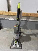 Full view of Hoover upright vacuum cleaner showing canister style, gray body with lime-green accents, clear dust container, and flexible hose.