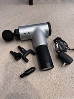 Photo of a silver and black handheld massage gun with four black massage attachments laid out on beige carpet next to a black power cord. The massage gun has a round ball attachment connected. Attachments include a round ball, forked, flat, and bullet-shaped head. Power cord included for charging.