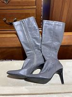 Side view of the pair of grey leather knee-high boots with visible side zippers and stiletto heels.