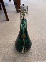 Tall green-tinted decorative oil candle bottle standing upright on carpeted floor. Bottle tapers at the top with a metal wick holder and hanging decorative chain with a tassel and medallion.