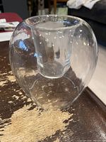 Clear glass part light candle/tea light holder placed on a wooden surface with visible scratches. Rounded outer shape with inner cup for candle placement, clean and transparent glass.