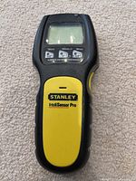 Black and yellow Stanley IntelliSensor Pro stud finder on carpeted floor showing the front with LCD screen and buttons.