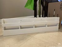 White wall mounted magazine rack placed on the floor, showing front side with three compartments and horizontal slats.
