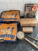 Photo showing two older blue and beige General Electric Movie Light boxes, vintage A E N Decorating Set box, a Maitland Charts Limited box, dry pencil, round metal container, and a vintage Maple Leaf magazine.