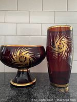 Photo showing ruby red footed bowl and tall vase with gold decorative designs on black counter and white tile background