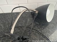 Black desktop clamp lamp showing conical shade, flexible silver metal neck, and clip clamp base on a granite countertop with white tiled backsplash.