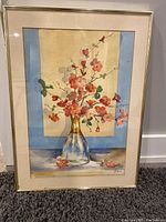 Framed watercolor painting showing a floral arrangement of pink and red blossoms in a glass vase, placed on a table with a beige and blue background.