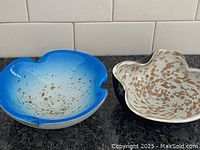 One photo showing the two MCM art glass ashtrays side by side on a dark granite surface. The blue ashtray has brown speckles and a visible hairline crack. The cream ashtray has brown speckles as well.