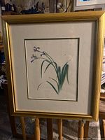 Framed botanical floral art with gold frame and cream mat, depicting green leaves and small purple flowers.