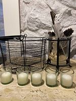 Five glass candle holders with white candles, a black wire basket, and a glass container with natural decorations inside a black metal frame arranged on a tabletop in front of a white textured wall.