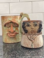 Two signed pottery mugs featuring artistic face designs, shown together