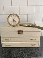Photo showing Westclox Baby Ben alarm clock sitting on top of cream jewelry box, gold hand mirror lying flat.