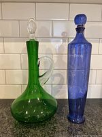 Front view of green glass decanter with clear stopper and blue glass vase with round stopper against white tile background