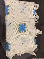 Folded view of the crocheted blanket showing cream base color with blue and green floral motifs and fringe.