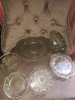 Photo showing six vintage glass/crystal pieces with intricate pressed patterns on a tufted chair.