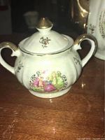 Close-up of sugar bowl with lid showing gold-trimmed handles and classical painted scene with figures and floral detail.