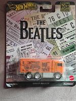 Front view of Hot Wheels collectible toy truck in packaging themed for The Beatles. Truck is orange with Beatles-themed graphics and is sealed inside original packaging with ticket stub designs.