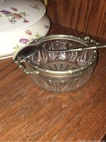 Clear glass bowl with silver-tone rim and handles, showing intricate cut-glass patterns, and a silver plate spoon resting inside.