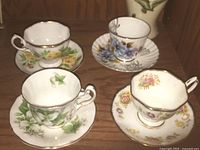 Photo showing the four teacup and saucer sets arranged on a wooden surface in natural lighting, highlighting floral patterns and condition.