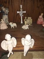 Image showing the two white angel figurines sitting at edge of shelf with other figurines including lambs, picture frame, cross in background.