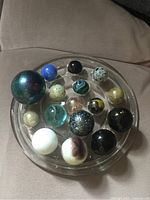 Photo showing 17 assorted marbles arranged on a clear glass dish along with a large blue-green speckled glass frog figurine.