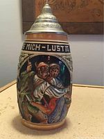 Front view of the beer stein showing colorful depiction of two people in traditional clothing and part of the raised text band near the top.