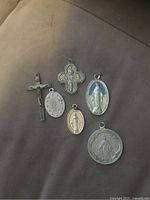 Photo showing six religious pendants including crucifix and Virgin Mary medals arranged on a flat surface.